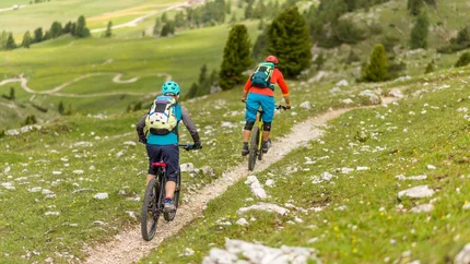 The Great Fanes Empire © Kirsten Sörries Two mountain bikers riding on a narrow trail through green mountainous terrain