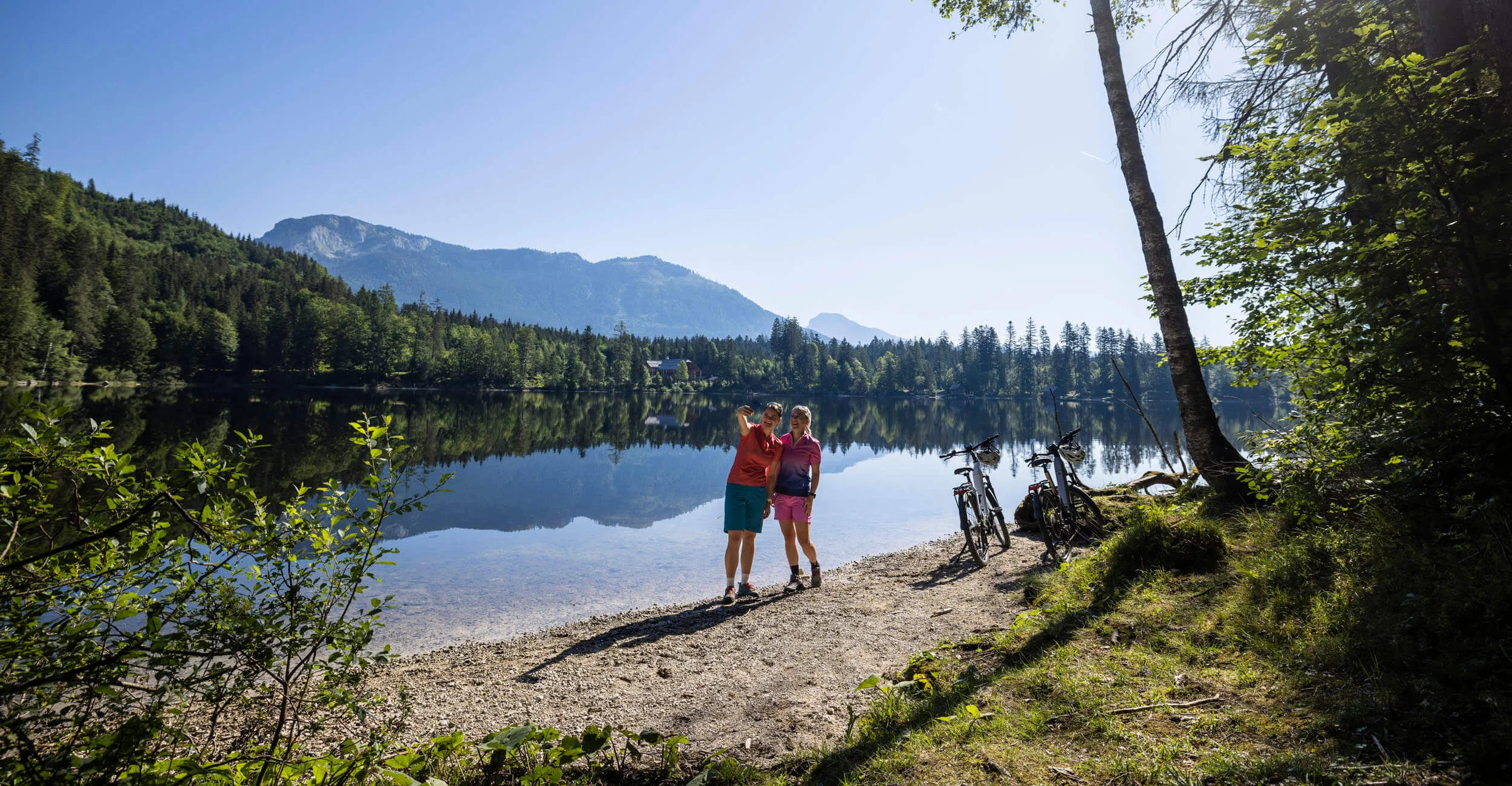 Multi-day tours © Velontour Two cyclists taking a selfie by the lake with mountains in the background