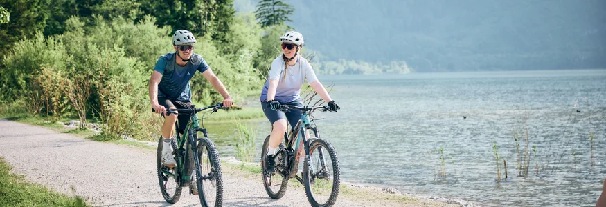 Holiday Region Dachstein Salzkammergut © Tobias Köhler Two cyclists riding bikes on a lakeside path with trees in the background