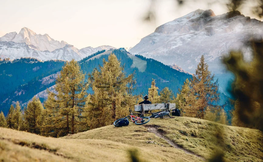 Mountain bike holidays in Alta Badia © Tobias Köhler Person sitting on bench in mountains with two mountain bikes and forest behind