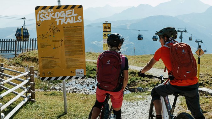 Mountain Bike Holidays © Felix Saller Zwei Mountainbiker lesen ein Schild an den Isskogel Trails mit Seilbahn im Hintergrund