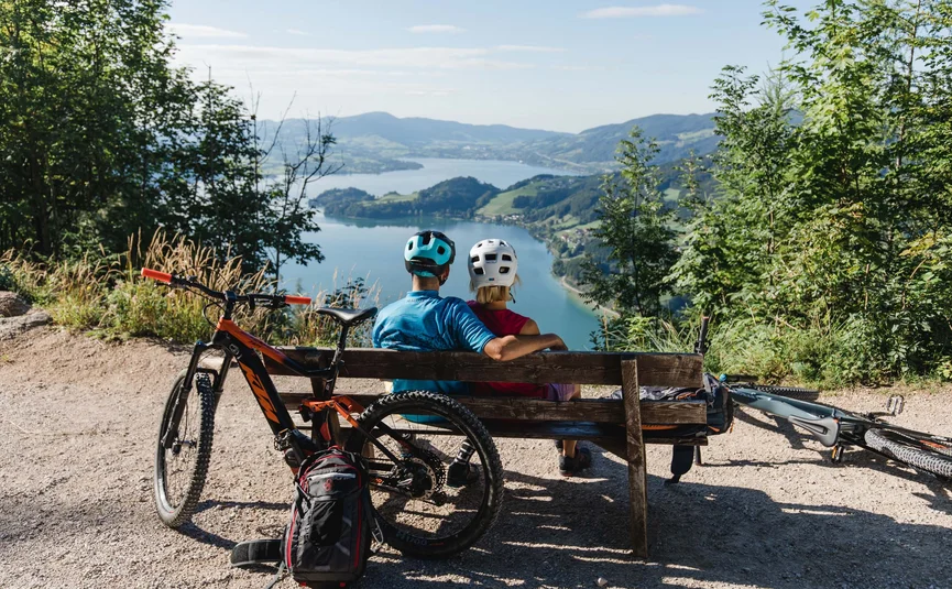 Mountain bike holiday in the Salzkammergut © Katrin Kerschbaumer Two cyclists sitting on a bench overlooking a lake and mountains.