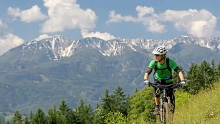 PODPECA - PECA TOUR © Urosh Modlic Mountain biker in green rides on trail with forested hills and snowy Alps in background