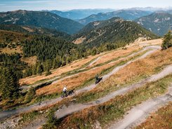 NOCKBIKE region © Jens Scheibe Two cyclists on a winding mountain trail surrounded by forests and mountains