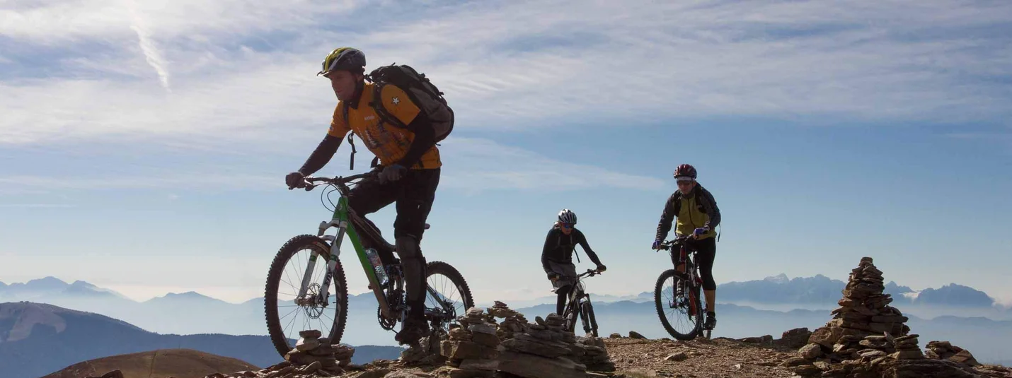 Alpe Adria World War Transalp © Hotel Lindenhof Three mountain bikers riding on a rocky mountain trail under a clear sky