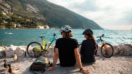 Trentino © David Karg Two cyclists wearing helmets sitting by a lake with mountains and water view