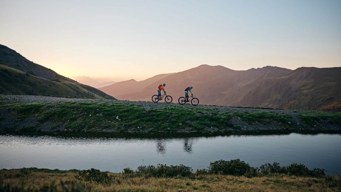 Mountain Bike Holidays © Tobias Köhler Zwei Mountainbiker fahren bei Sonnenuntergang auf einem Bergweg neben einem See