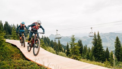 Kitzbühel Alps © Patrick Wasshuber Two mountain bikers riding downhill on a wooden trail surrounded by pine trees