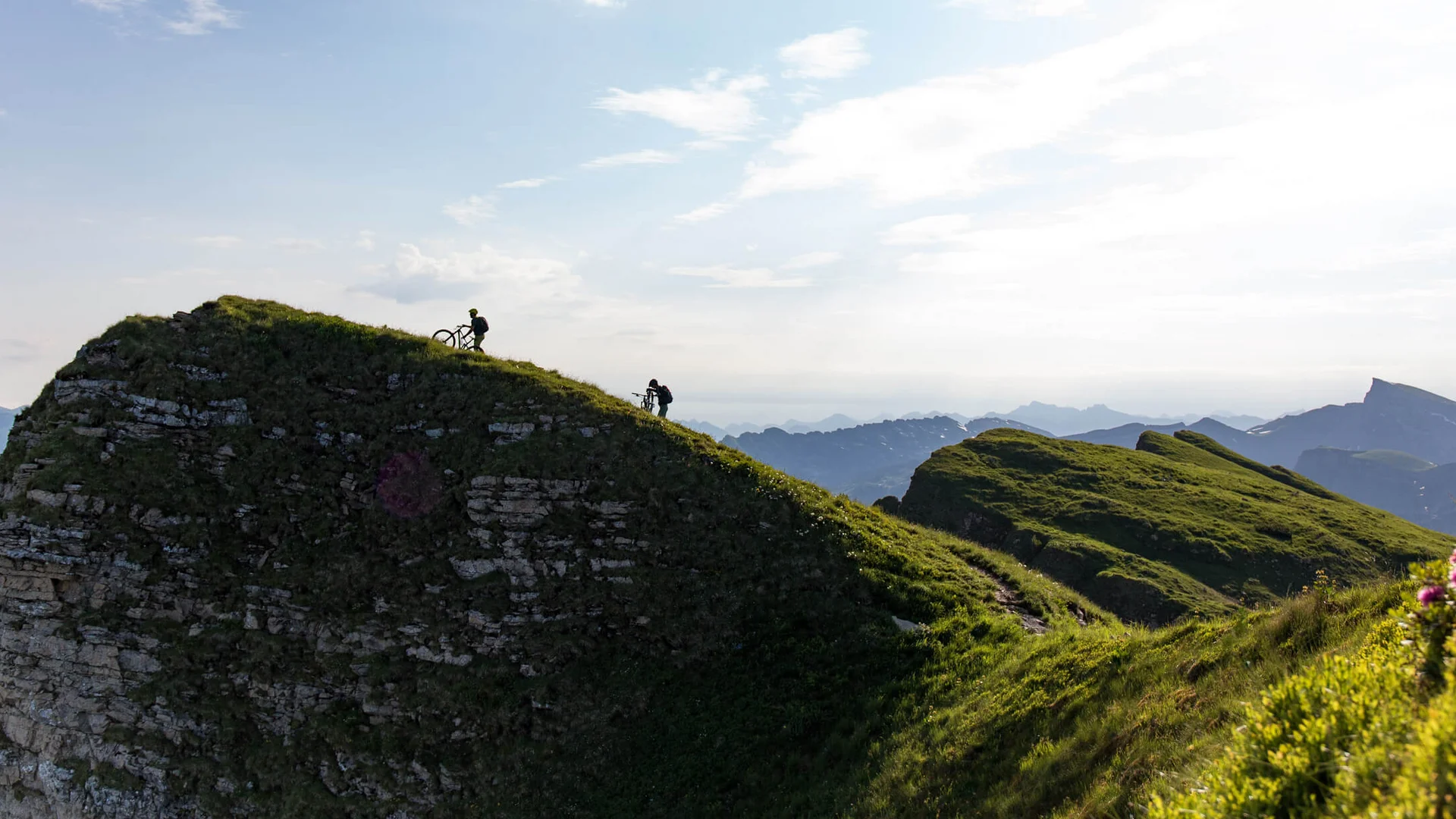 Diedamskopftrail © David Karg Two cyclists pushing bikes up grassy hill in mountainous landscape