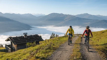 RAUHER KOPF © TVB Kitzbüheler Alpen Brixental - Mathäus Gartner Two cyclists riding on a mountain trail with fog in the valley and mountains