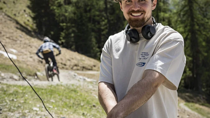 Mountain Bike Holidays © Manfred Stromberg Lächelnder DJ mit Kopfhörern draußen vor Bäumen und Radfahrer im Hintergrund