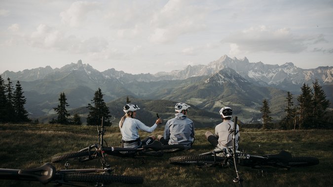 Mountain Bike Holidays © Matthias Warter Drei Radfahrer sitzen auf einer Wiese mit Mountainbikes vor Berglandschaft