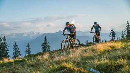 Rossbrand - Mandling Tour Three mountain bikers cycling on a grassy hill with mountains in the background
