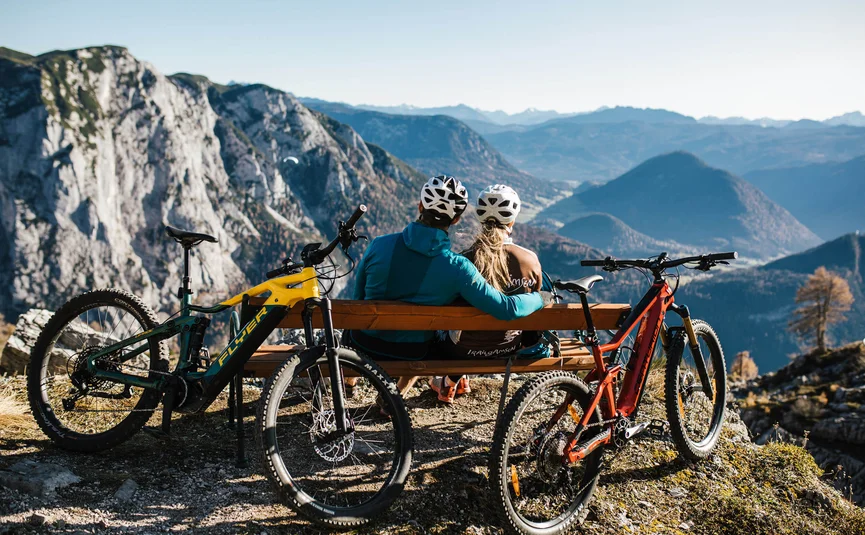 Mountain bike holidays in Styria © TVB Ausseerland Salzkammergut - Katrin Kerschbaumer Two cyclists sitting on bench overlooking mountain landscape with two mountain bikes