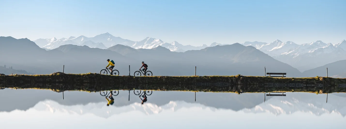 SAUKASER - PENGELSTEIN © TVB Kitzbüheler Alpen Brixental - Mathäus Gartner Two cyclists riding by a lake with mountain reflection and clear blue sky