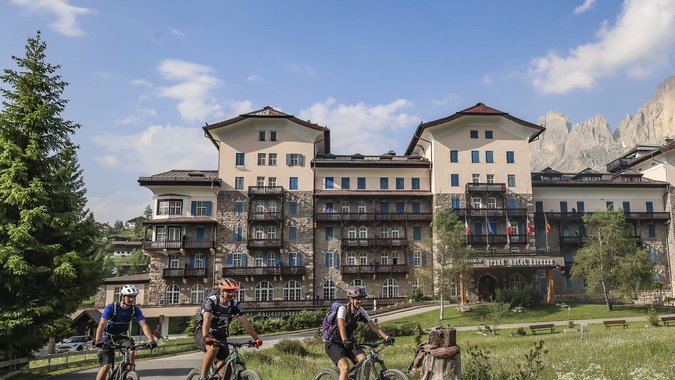 Mountain Bike Holidays © Jens Vögele - Westsiders Drei Radfahrer vor einem großen Berghotel mit Bergen im Hintergrund