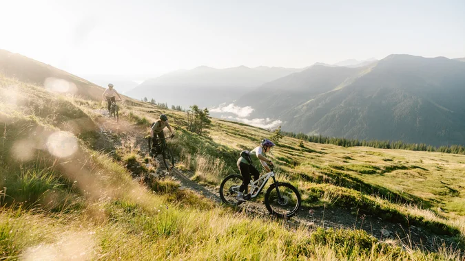 Mountain Bike Holidays © saalbach.com, Moritz Ablinger Drei Mountainbiker fahren auf einem Bergpfad im Sonnenlicht