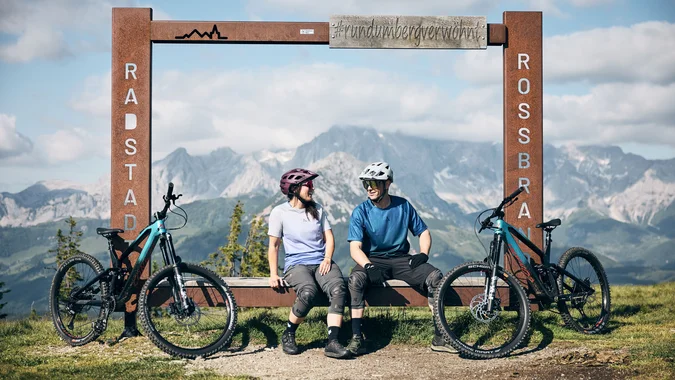 Mountain Bike Holidays © Tobias Köhler Zwei Radfahrer sitzen neben ihren Fahrrädern an einem Bergblick-Rahmen in Radstadt Rossbrand.
