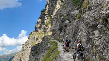 Around the Mittagsfluh © Alpen Hotel Post Two cyclists push mountain bikes on a narrow mountain trail along a steep rock face