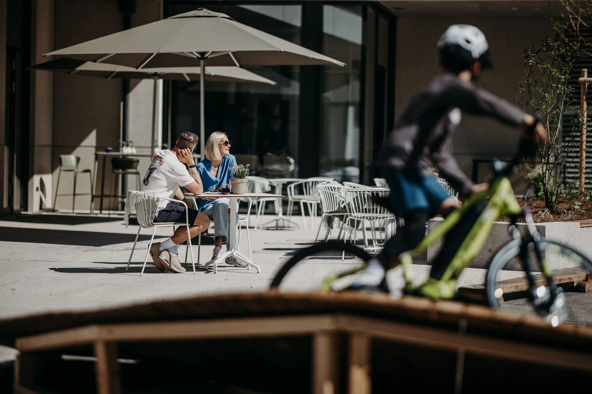 Mountain Bike Holidays © Matthias Warter Paar sitzt draußen unter Sonnenschirmen, Radfahrer fährt vorbei