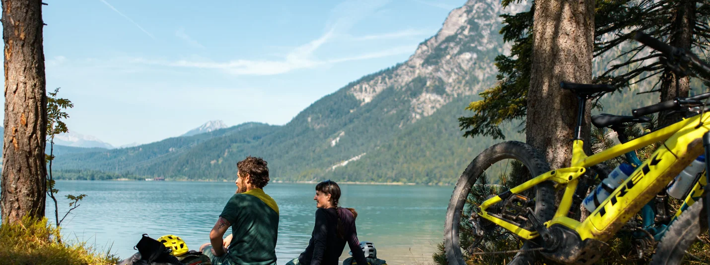FROM SCHÖNRUH TO LAKE SEEBENSEE © David Karg Two cyclists sitting by a lake with mountains in the background
