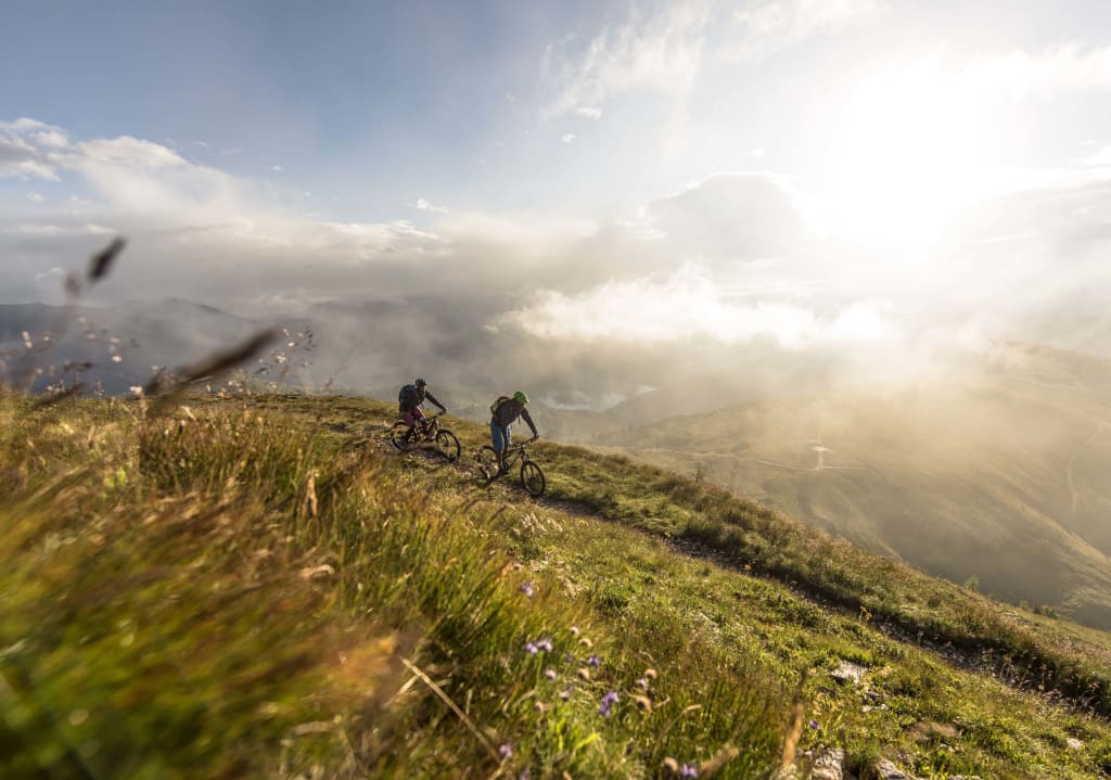 Bikedays Early Bird -10% discount Two mountain bikers riding on a mountain trail with sunlight and fog