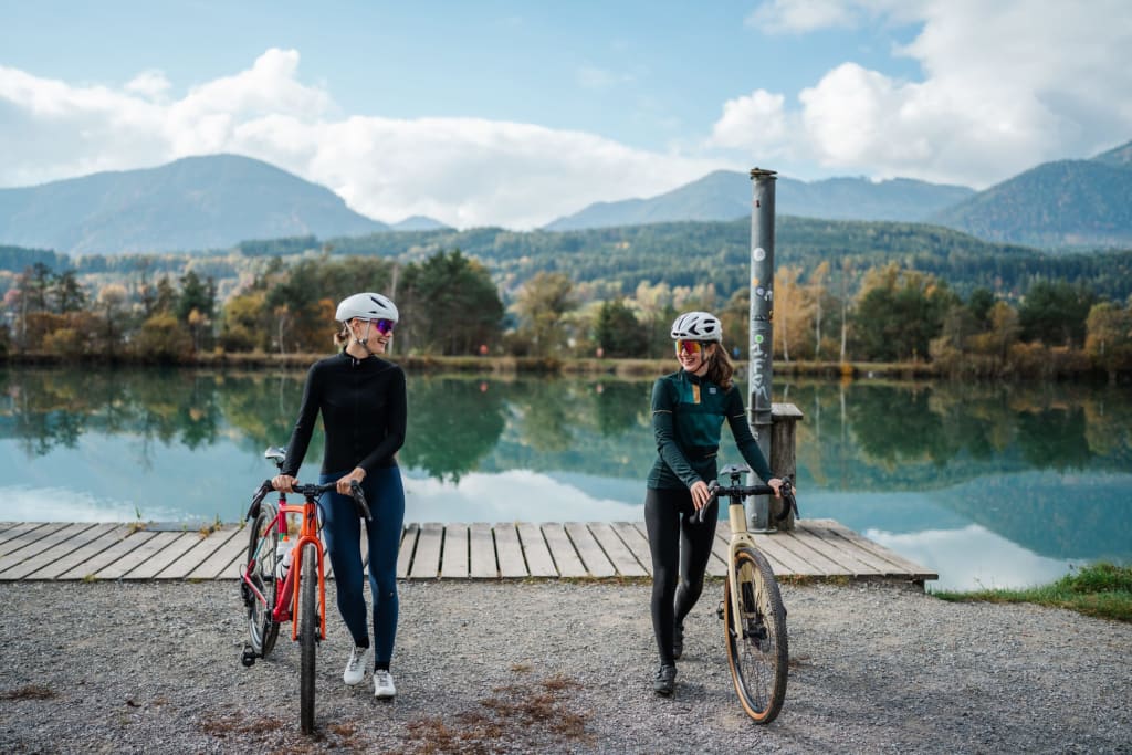 Gravel & Glory Two female cyclists with bikes by lake shore with mountains in background